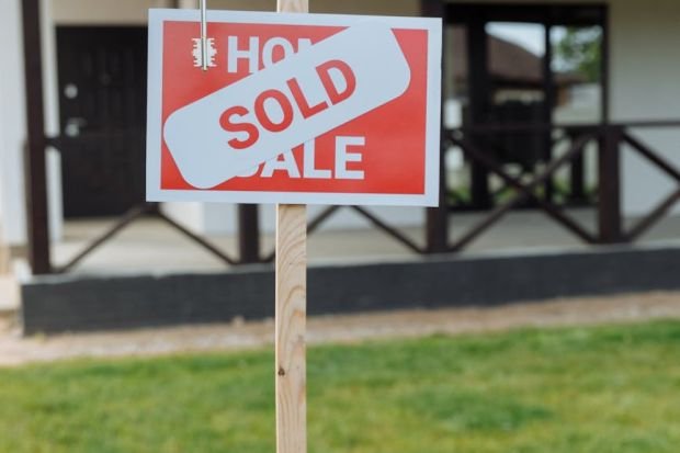 A hand holding a key in front of a sold sign and a house, symbolizing a successful real estate transaction.