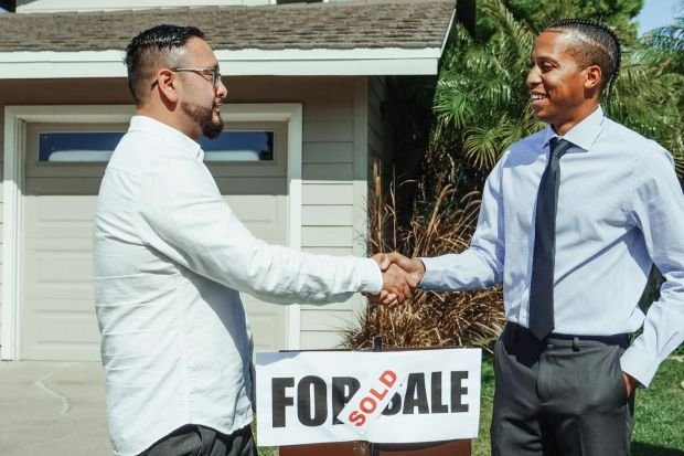 Two men shaking hands in front of house sold sign, sealing real estate deal outdoors.