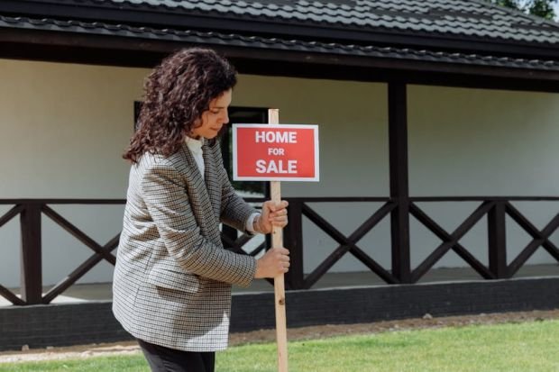 Woman real estate agent placing a 'Home for Sale' sign on a green lawn in front of a house.
