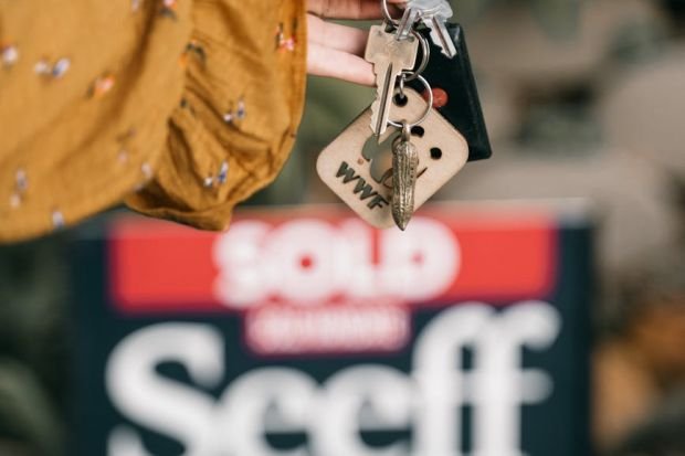 A hand holds keys in front of a blurred sold real estate sign, symbolizing completed sale.